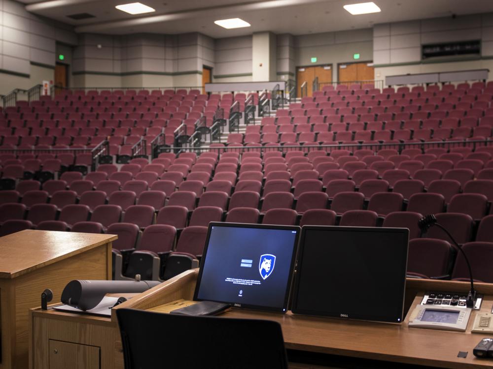A computer screen is lit up at a podium overlooking an empty theater classroom.